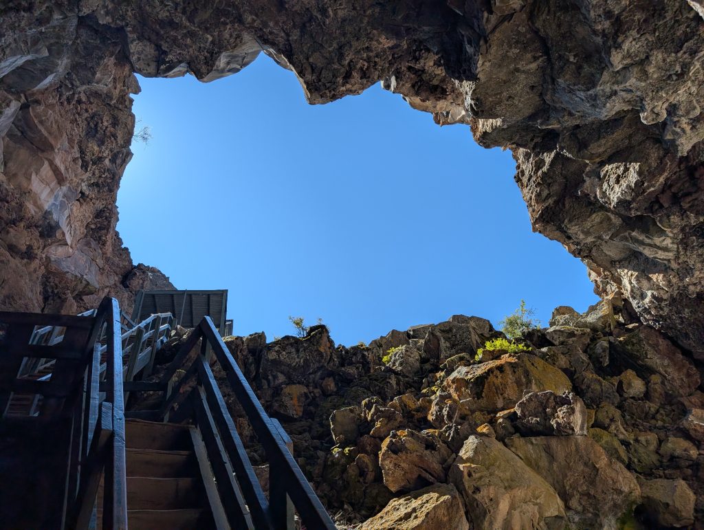 A low-angle shot from the bottom of the Bandera Ice Cave in New Mexico, looking up a steep, dark wooden staircase toward a circular opening of bright blue sky.